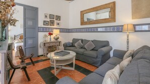 Een woonkamer met een grijze bank, een ronde glazen salontafel en een houten stoel. Er staat een kleine tafel met een lamp en een vaas met bloemen. De kamer heeft een betegelde muur en een spiegel.