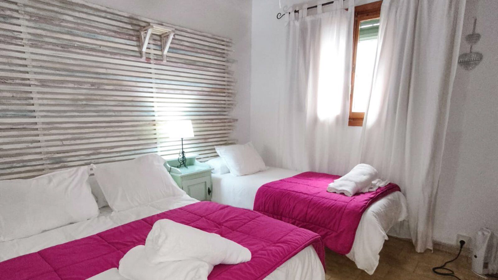 A hotel room with two beds, a window with white curtains, a bedside table with a lamp, and a wall-mounted air conditioning unit.