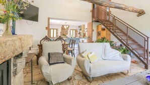 Un salon chaleureux avec une cheminée en pierre, un escalier en bois et, au fond, un coin repas.