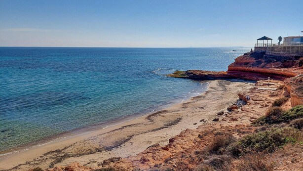 Uma praia costeira com águas azuis cristalinas, falésias rochosas e uma pequena construção à direita.