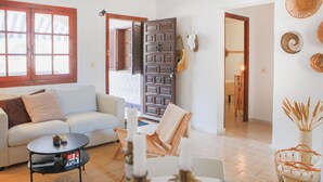 Sala de estar com um sofá de cor clara, uma mesa de centro redonda e uma cadeira de vime. Há uma porta de madeira com olho de fechadura, um porta-chapéus fixado na parede e uma cesta de vime no chão.