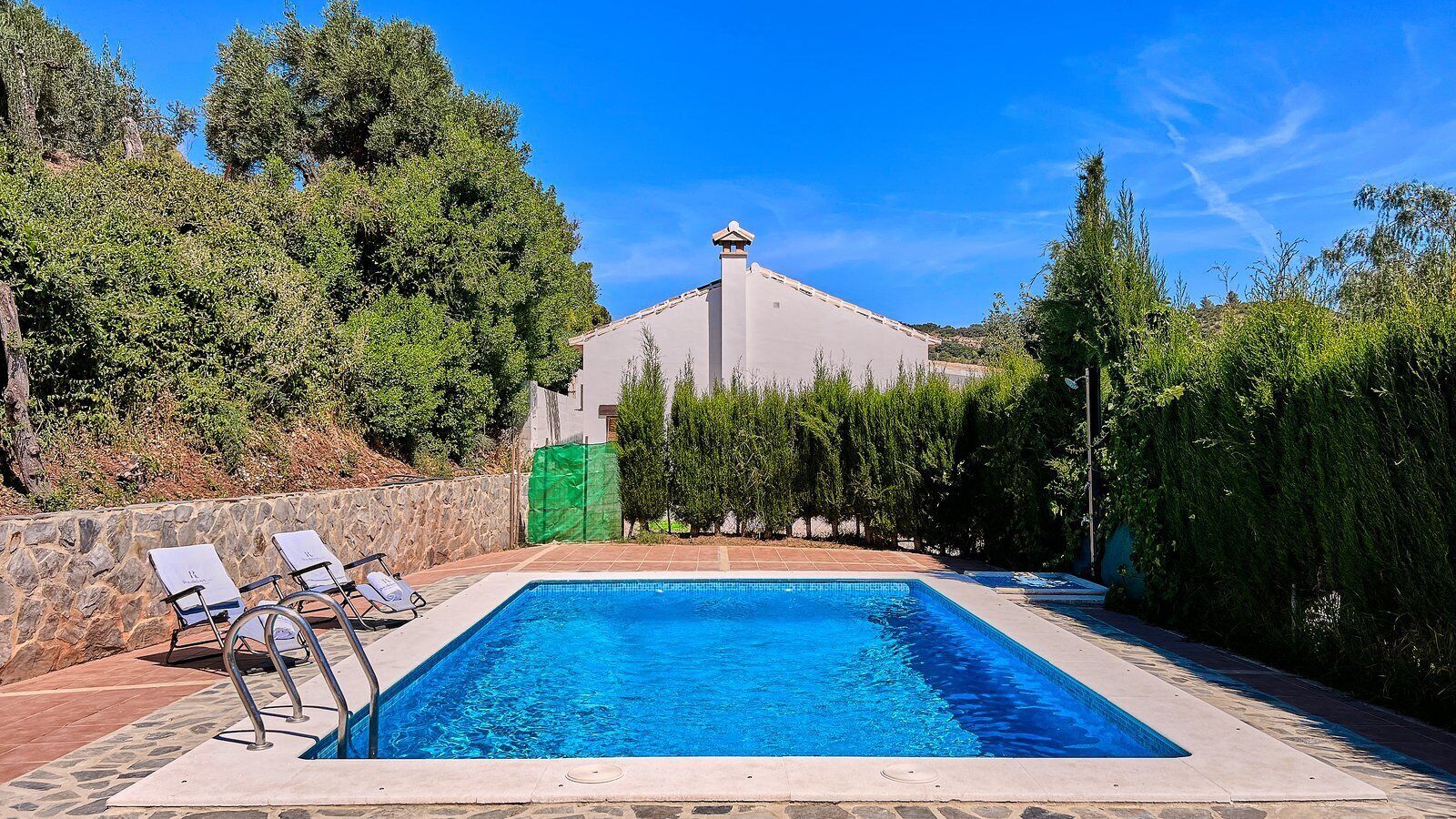 Une piscine rectangulaire à l’eau d’un bleu cristallin, entourée d’un mur de pierre et bordée d’une rangée de chaises longues blanches.