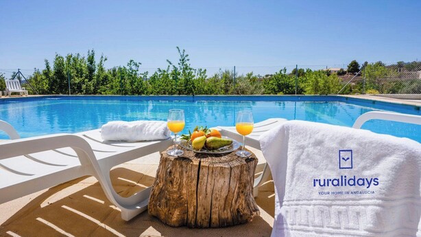 Uma área na beira da piscina com um toco de madeira, uma espreguiçadeira branca, uma toalha e um prato de frutas com bebidas.
