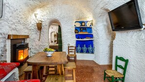 A rustic interior with a stone wall, a wood-burning stove, a wooden table with chairs, a TV mounted on the wall, and decorative blue and white items. - La Cueva de la Abuela  Cenes de la Vega by Ruralidays (Granada)