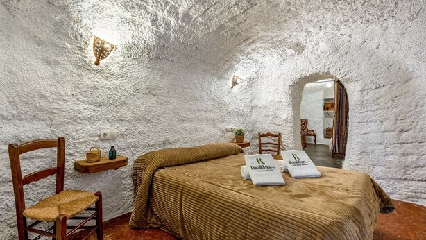 A rustic bedroom with a wooden bed, a chair, a shelf with a lamp, and towels on the bed. - La Cueva de la Abuela  Cenes de la Vega by Ruralidays (Granada)