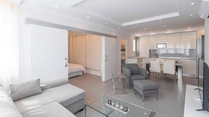 A modern living room with a sofa, ottoman, and glass coffee table. A kitchen area with a dining set is visible in the background. - Lux Playa Fuengirola by Ruralidays (Málaga)