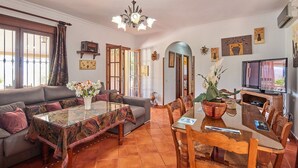 Ein Wohnzimmer mit einer Couch, einem Glastisch, einem Esstisch mit Stühlen, einem Fernseher und verschiedenen Wanddekorationen.