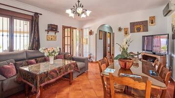 Una sala de estar con sofá, una mesa de centro de vidrio, una mesa de comedor con sillas, un televisor y varias decoraciones en las paredes.