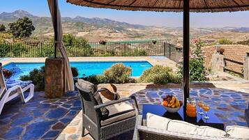 Zona de piscina con vista a montañas, piso de piedra, sillas de descanso, una silla de mimbre, una mesa con frutas y bebidas, y una pérgola.