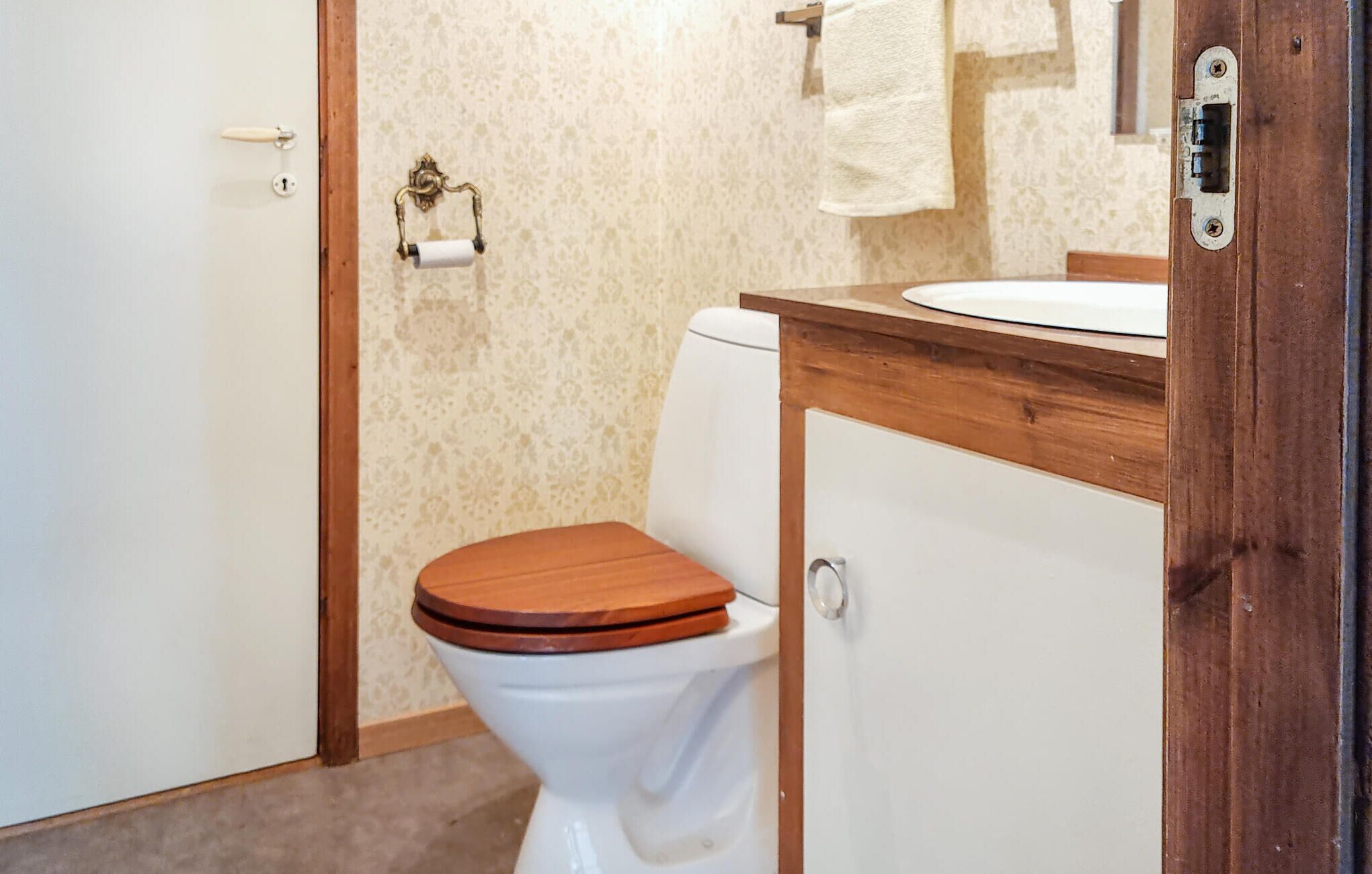 Un baño con inodoro, tocador de madera, toalla colgada en un perchero y puerta con cerradura.