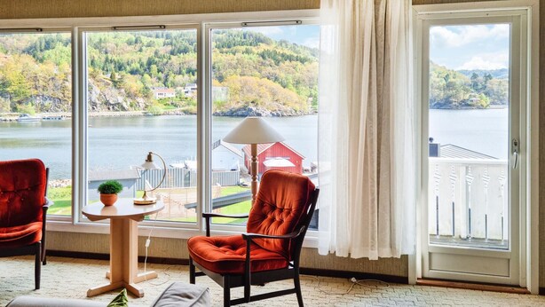 Ein Zimmer mit großem Fenster mit Blick auf einen See und Wald, ein roter Sessel, ein kleiner Tisch mit Lampe sowie ein Blick auf ein rotes Gebäude gegenüber dem Wasser.