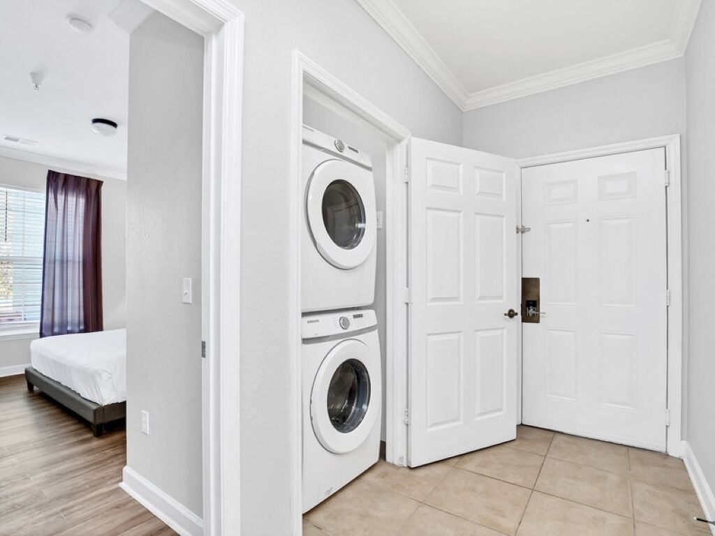 A laundry room with a washing machine and dryer, a door, and a bed in the adjacent room.