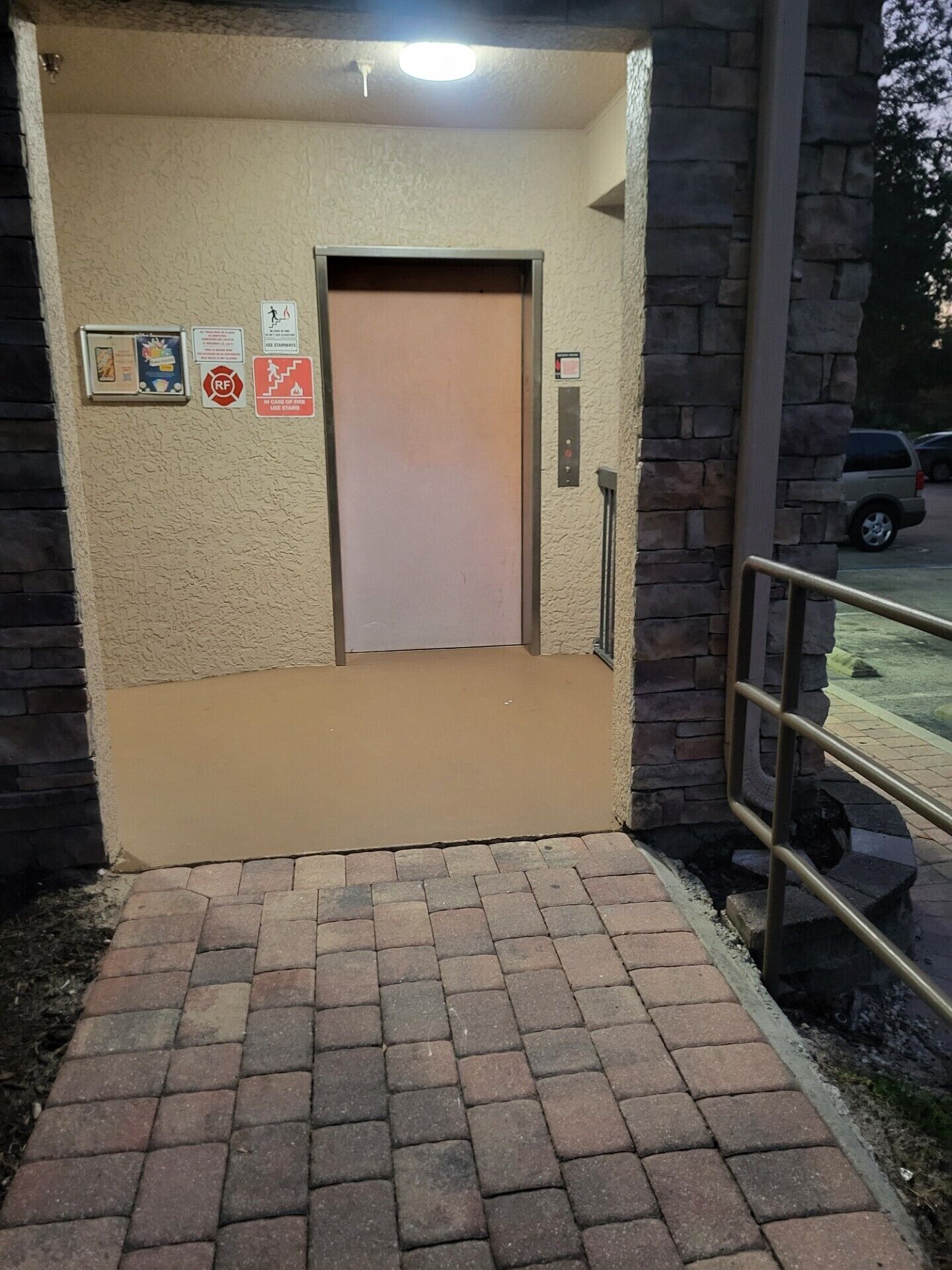 An elevator entrance with a beige wall, a metal handrail, and a brick pathway leading to it.