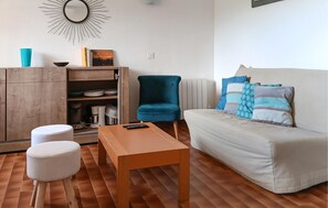 A living room with a wooden table, a blue armchair, a white sofa with patterned pillows, a wooden cabinet with kitchen items, and a wall-mounted mirror.