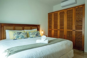 A neatly made bed with a headboard, a set of wooden louvered closets, and a wall-mounted air conditioner.