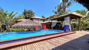 Une terrasse en bois avec une piscine, des constructions à toits de chaume et une végétation tropicale.