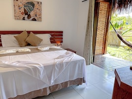 Una cama bien hecha con sábanas blancas y dos almohadas, un cabecero de madera, una mesita de noche con una lámpara, y vista a un jardín exuberante a través de una puerta abierta.
