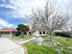 A two-story house with a red-tiled roof, a garage, and a well-maintained garden with a large tree and rocks. - Design newly remodeled  villa. 10 minutes from Palm Springs (Desert Hot Springs)