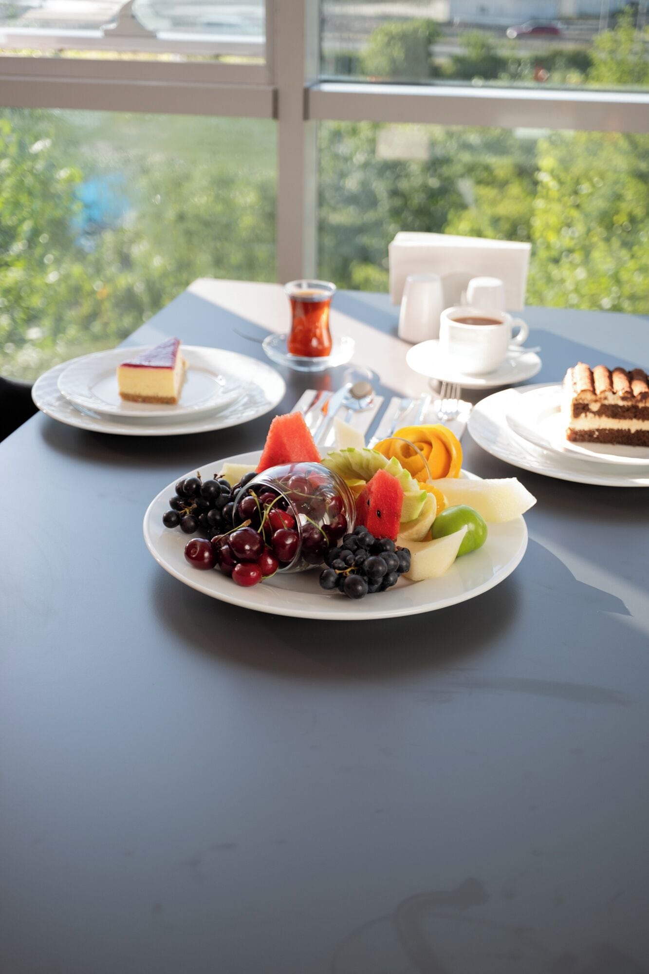 A table with plates of fruit and desserts, cups of coffee, and a glass of tea.
