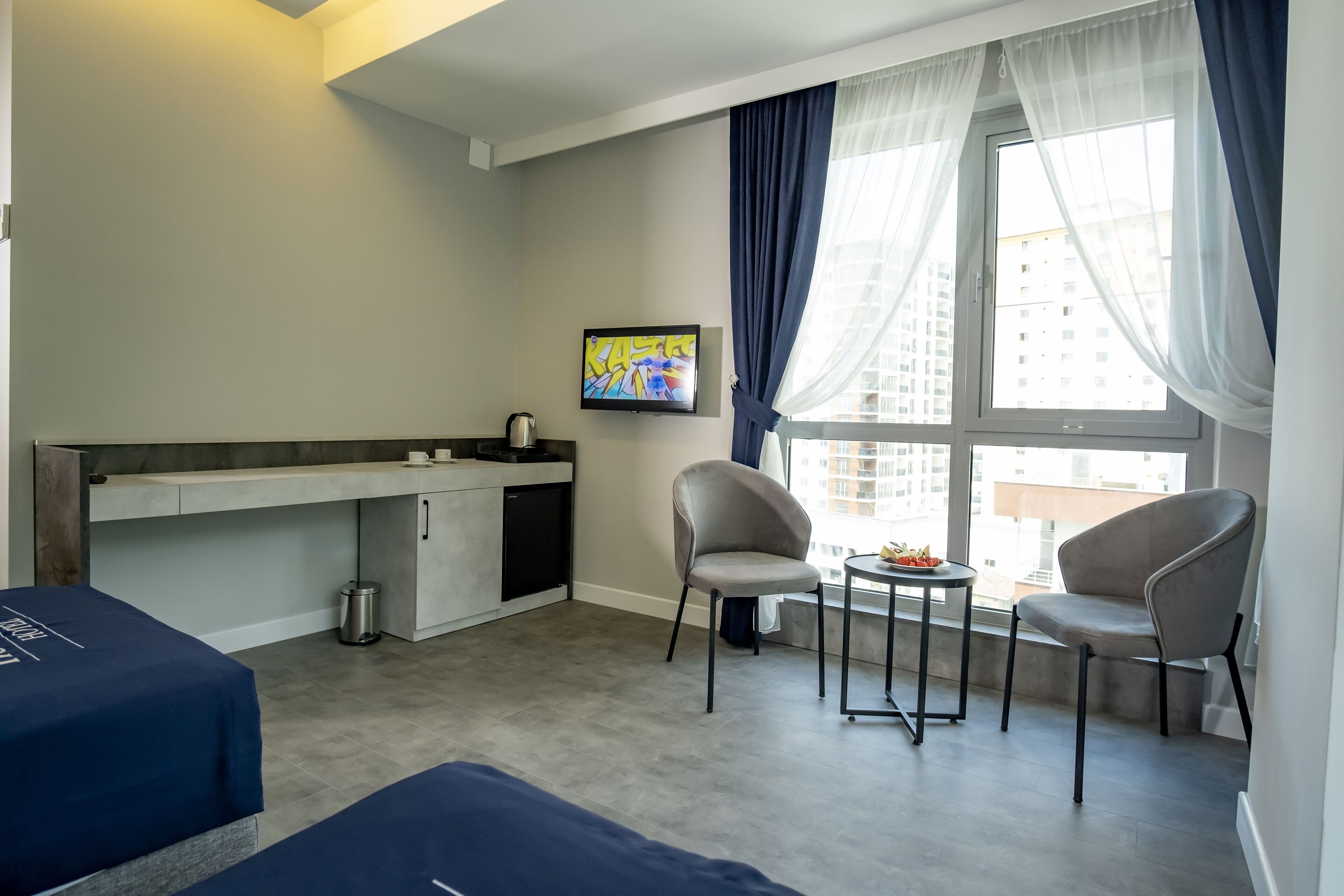 A compact hotel room with a kitchenette, two chairs, a small table, and a large window with curtains.