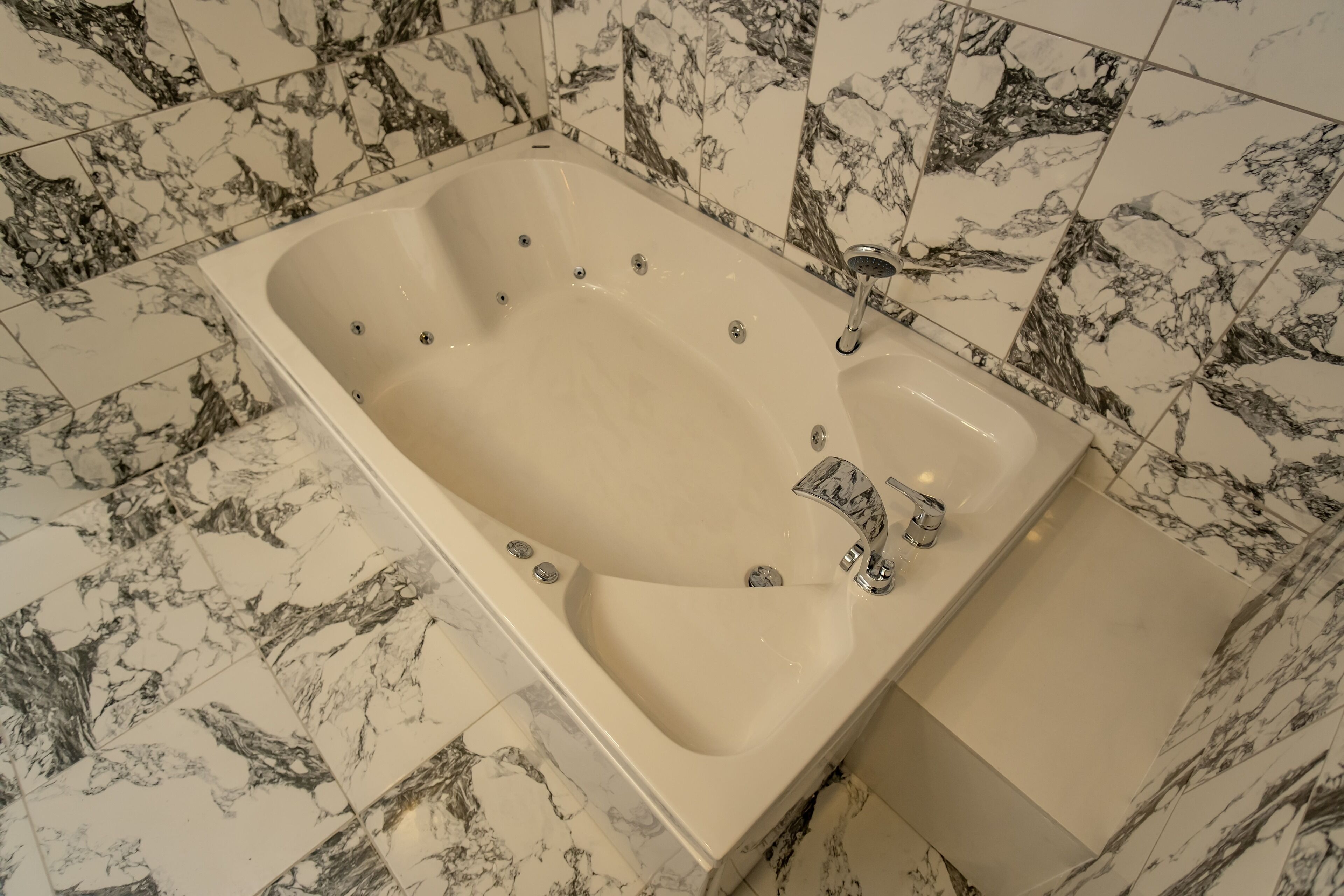 Una bañera rectangular con un solo grifo y un cabezal de ducha manual, frente a un fondo de azulejos de mármol.