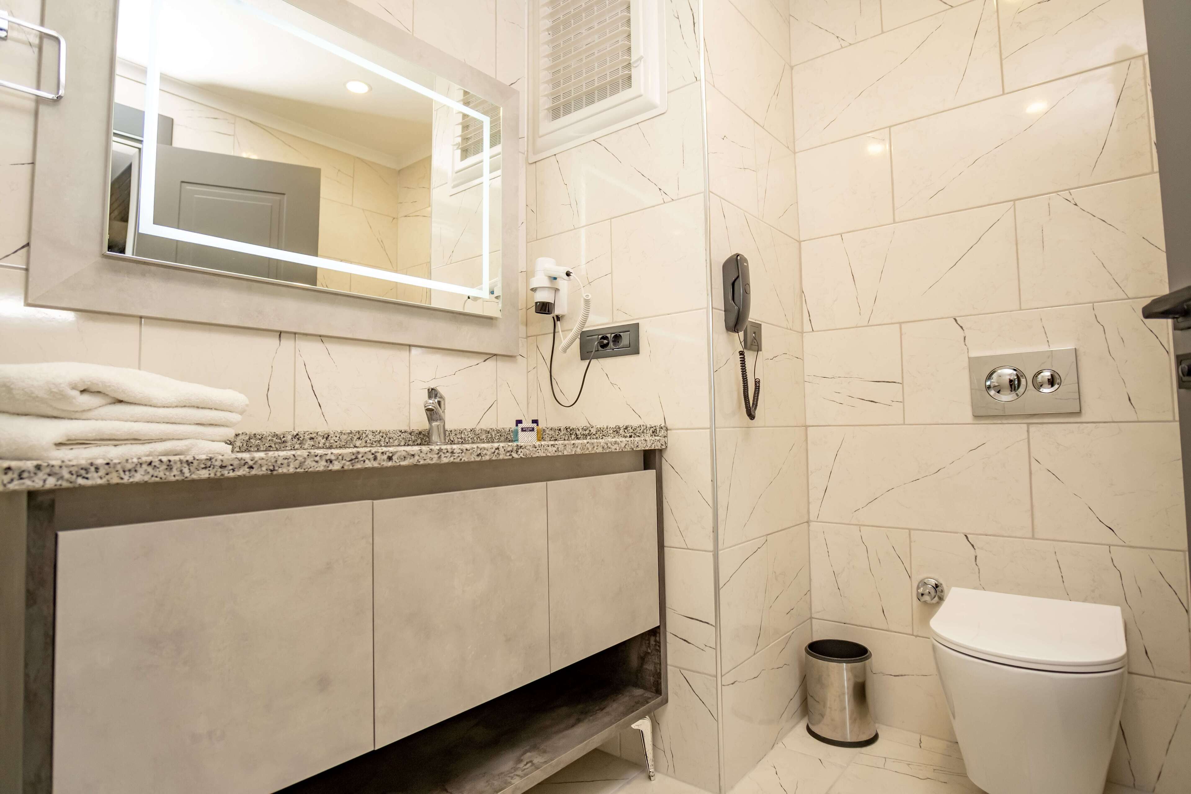 A modern bathroom with a marble-tiled wall, a vanity with a sink, a hairdryer, a telephone, and a toilet.