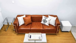 Ein braunes Ledersofa mit gemusterten Kissen, ein weißer Hocker und ein Glastisch mit Fernbedienung.