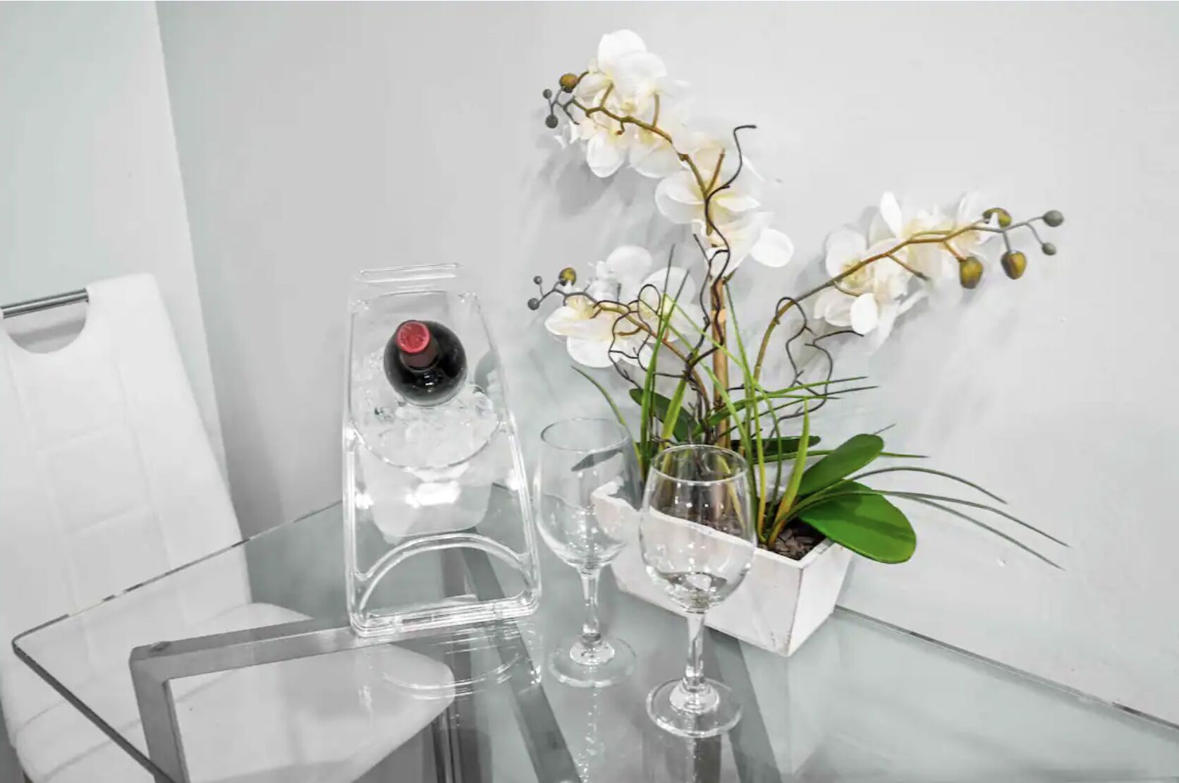 Une table en verre transparent sur laquelle se trouvent une bouteille de vin rouge, deux verres à vin et une plante d’orchidée blanche dans un pot blanc.