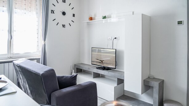 Un salon moderne avec un canapé gris, un téléviseur à écran plat posé sur une console blanche, une horloge murale et une fenêtre avec des rideaux.