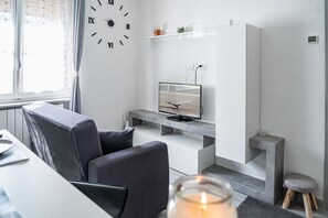 A modern living room with a grey sofa, a flat-screen TV on a white console, a wall clock, and a window with curtains.