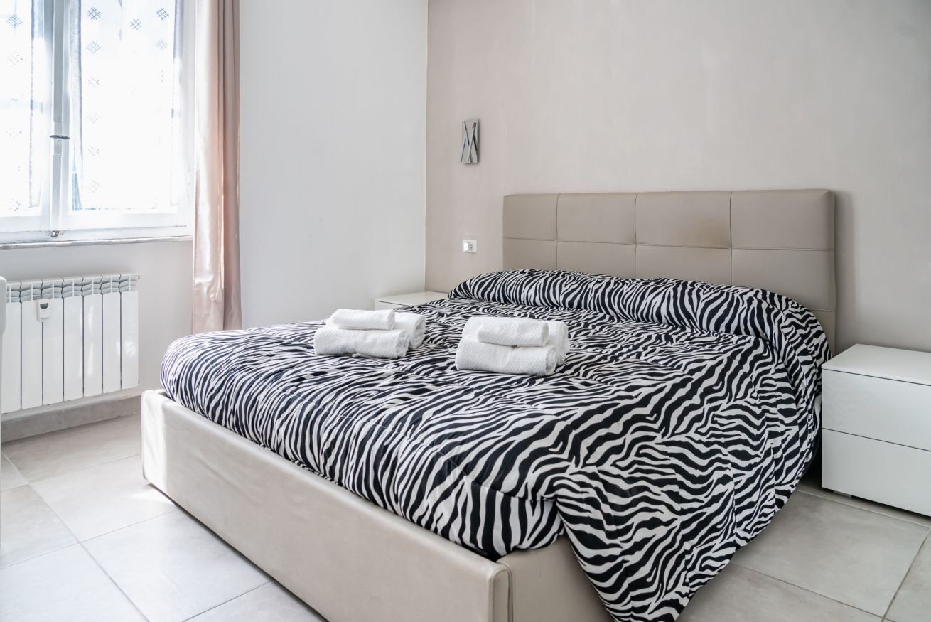 Une chambre à coucher avec une couvre-lit à motifs zébrés, une table de chevet blanche, un radiateur et une fenêtre avec des rideaux.