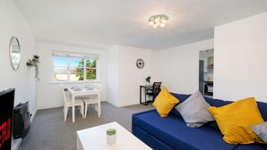 A modern living room with a blue sofa, white coffee table, and a TV. There is a dining area with a white table and chairs, and a window with a view of the outside. - 2 bed luxury apartment (Enfield)