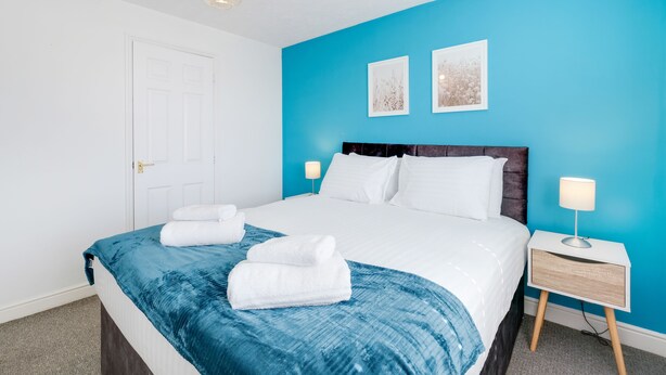 A neatly made bed with white linens and a blue accent throw, a bedside table with a lamp, and two framed pictures on the blue wall. - 2 bed luxury apartment (Enfield)