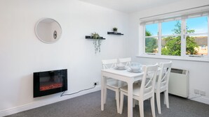 A dining area with a white table and chairs, a flat-screen TV, a fireplace, and a window with a view of trees and houses. - 2 bed luxury apartment (Enfield)