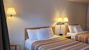 Ein Hotelzimmer mit zwei Betten, jeweils mit einem hölzernen Kopfteil und weißer Bettwäsche. An beiden Seiten der Betten befinden sich Wandlampen. In der Ecke ist ein Holzstuhl und ein kleiner Holztisch zu sehen.