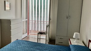 A bedroom with a bed, a wardrobe, a desk with a lamp, and a view of the outside through a door with a striped curtain.