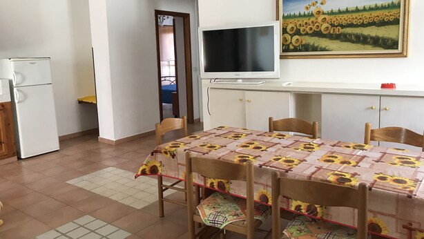 A dining area with a table covered in a sunflower tablecloth, wooden chairs, a television on a stand, and a painting of sunflowers on the wall.