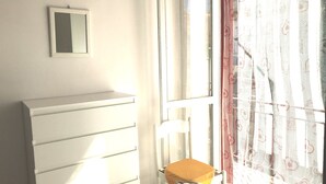 A room with a white dresser, a chair with a yellow cushion, a small mirror on the wall, and a window with patterned curtains.