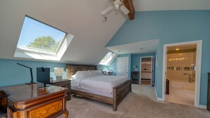 Une chambre avec un grand lit, un bureau et un ventilateur de plafond. Elle est dotée d’une lucarne et d’une porte donnant accès à une salle de bain.