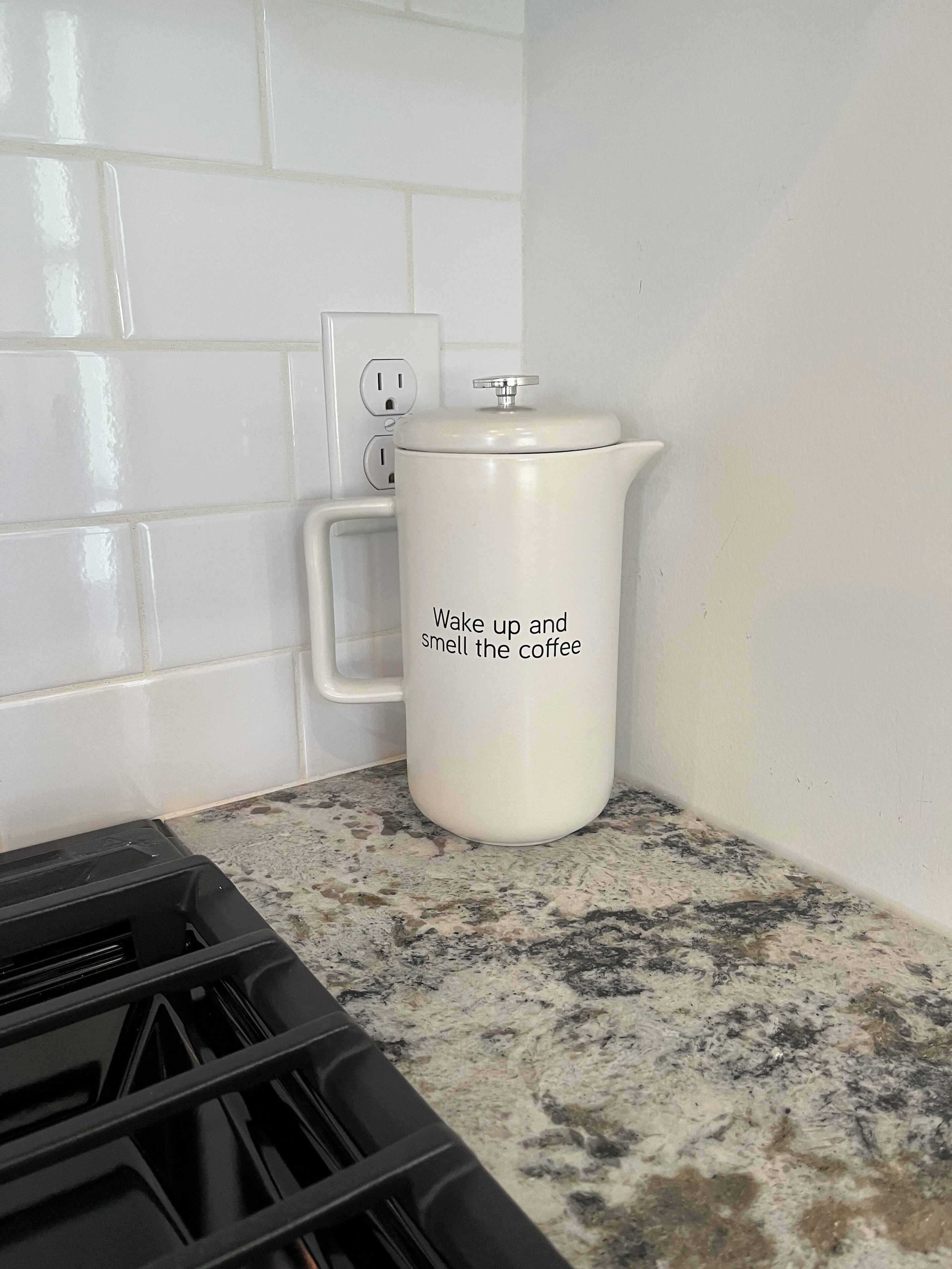 Una cocina con paredes de azulejos blancos, una cafetera blanca con tapa y una encimera de granito.