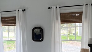 A bedroom with a bed, white curtains, a window with a view of a garden, a small mirror, and a lamp on a bedside table. - The Cottage At Hershey Lane Farm (Frenchtown)