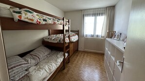 Kerrossängyllä varustettu huone, jossa on kalanruotokuviollinen lattia, patteri ja ikkuna, jossa on verhot.