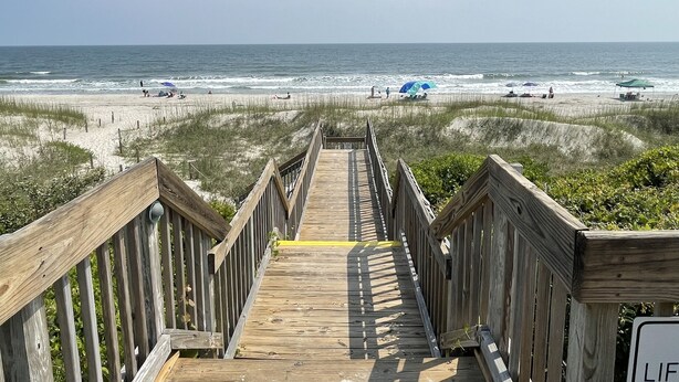 A wooden boardwalk leading to a beach with people and umbrellas. - Sea Turtle Sanctuary-Ocean Front Condo on Perfect Private Beach!  (Oak Island)