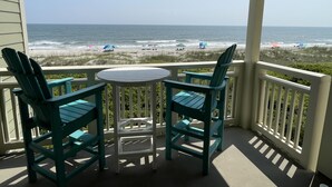A covered outdoor seating area with two chairs and a table, overlooking a beach with umbrellas and people. - Sea Turtle Sanctuary-Ocean Front Condo on Perfect Private Beach!  (Oak Island)