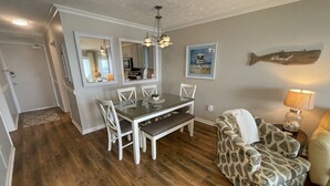 A dining area with a glass table and white chairs, a patterned sofa, and a wall-mounted fish decoration. - Sea Turtle Sanctuary-Ocean Front Condo on Perfect Private Beach!  (Oak Island)