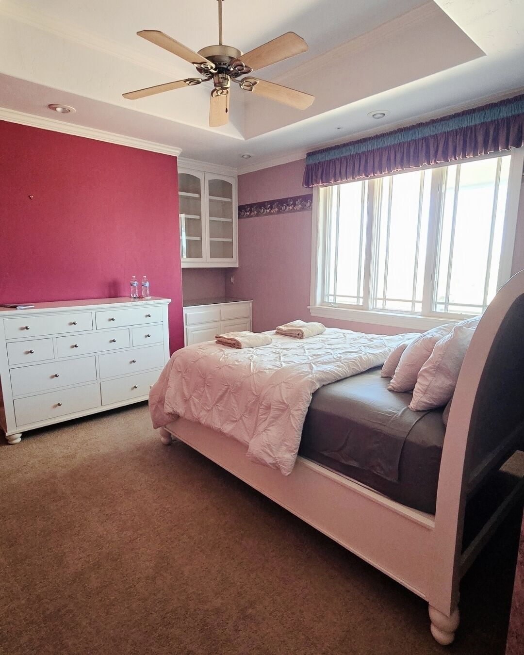 A bedroom with a bed, a dresser, a ceiling fan, and a window with curtains.