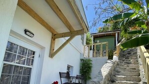 Una escalera de piedra lleva a una casa con una puerta blanca y una ventana. Cerca de la puerta hay una pequeña mesa con sillas y una planta en maceta.