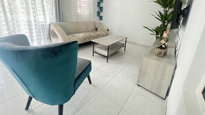 Una sala de estar con un sillón azul verdoso, un sofá color crema, una mesa de centro y un televisor sobre un mueble.