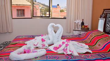 Habitación de hotel con una cama cubierta con un edredón colorido, una ventana con vistas a edificios y una mesita con una botella y fruta.