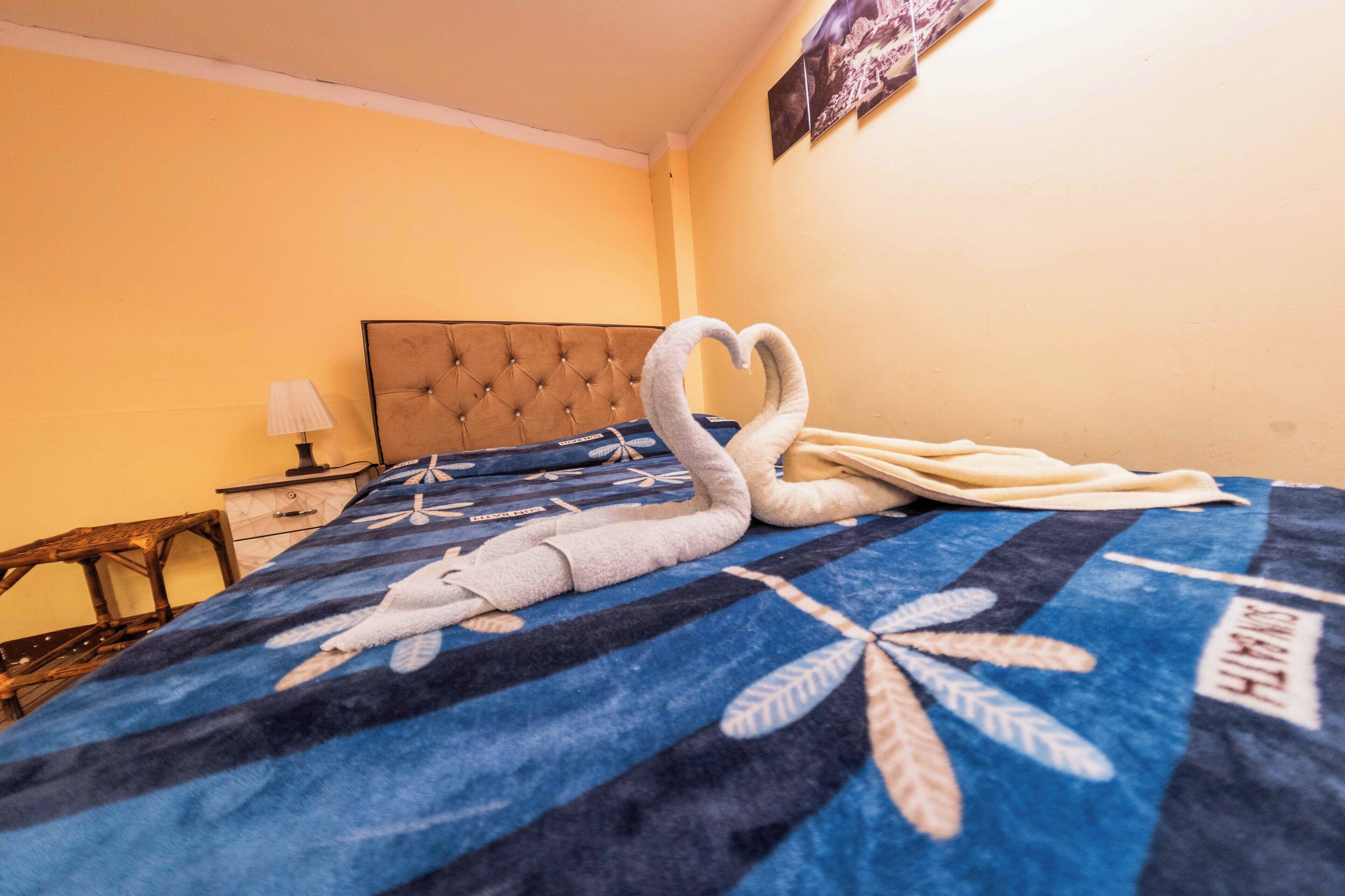 Habitación de hotel con una cama cubierta con una colcha azul y negra estampada, un cabecero capitonado, una mesita de noche con lámpara y un cuadro colgado en la pared.